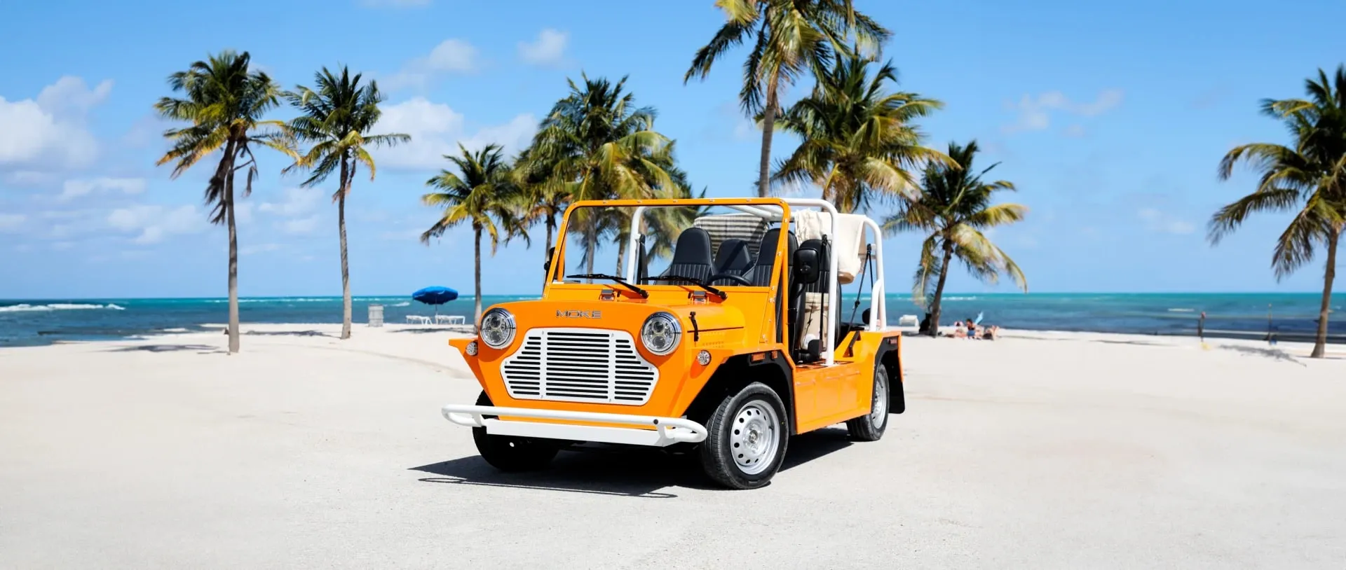 An orange Moke on white sand beneath palm trees